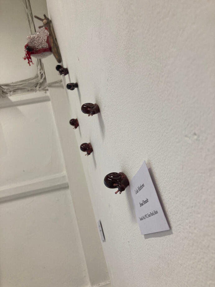 An angled image looking up at a needle felted snail with a shell full of pearls, which has red entrails of fabric hanging from it. Dark red resin snails are crawling down the wall away from the needle felted snail.
